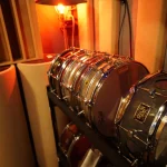 Full snare drum collection on rack under warm studio lighting at Somni Studios.
